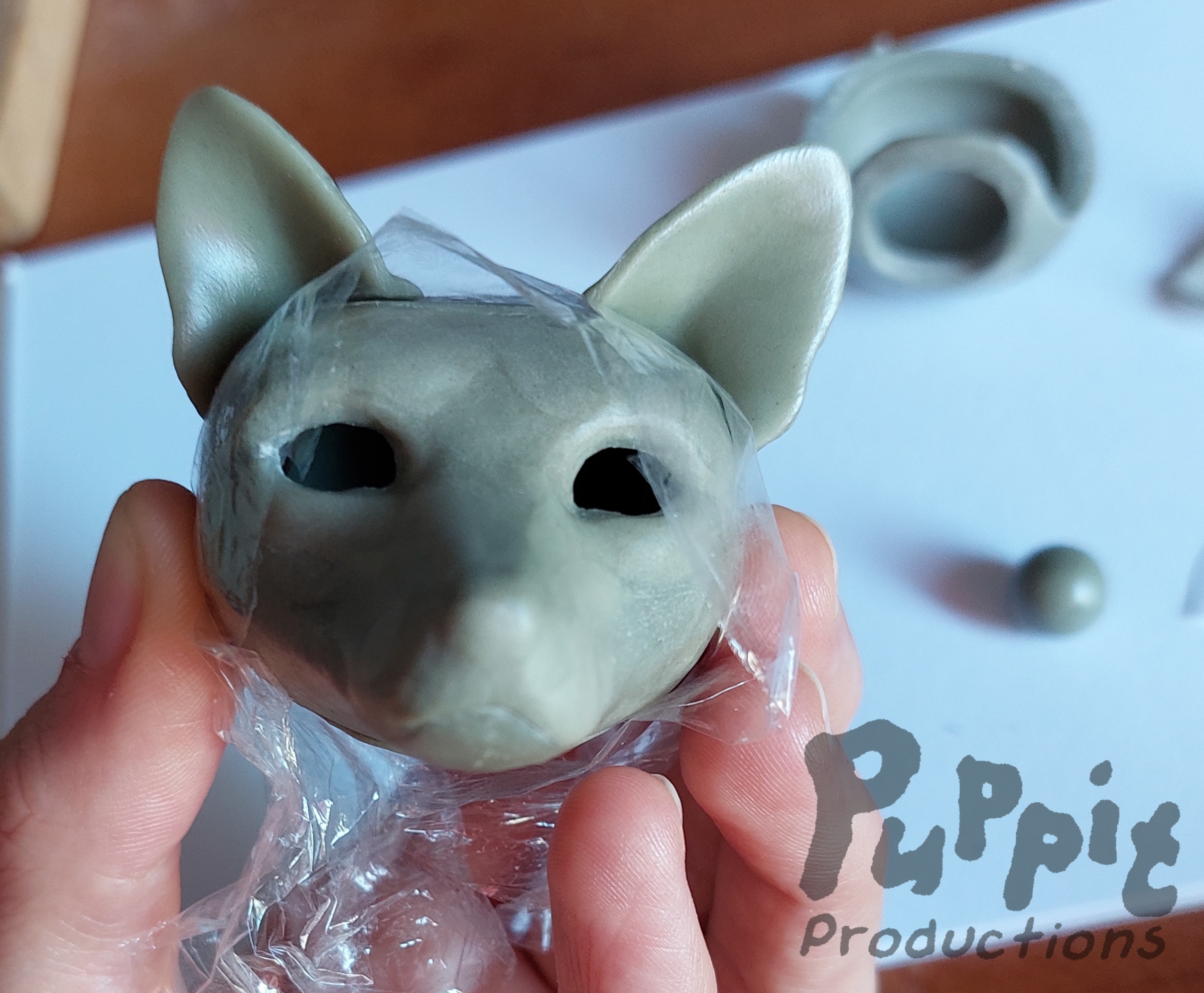 A picture of a red wolf head for a doll in rough gray clay finish, viewed from straight ahead. It does not have any eyes yet and between the ears and around the back of the head cellofane wrap is visible.