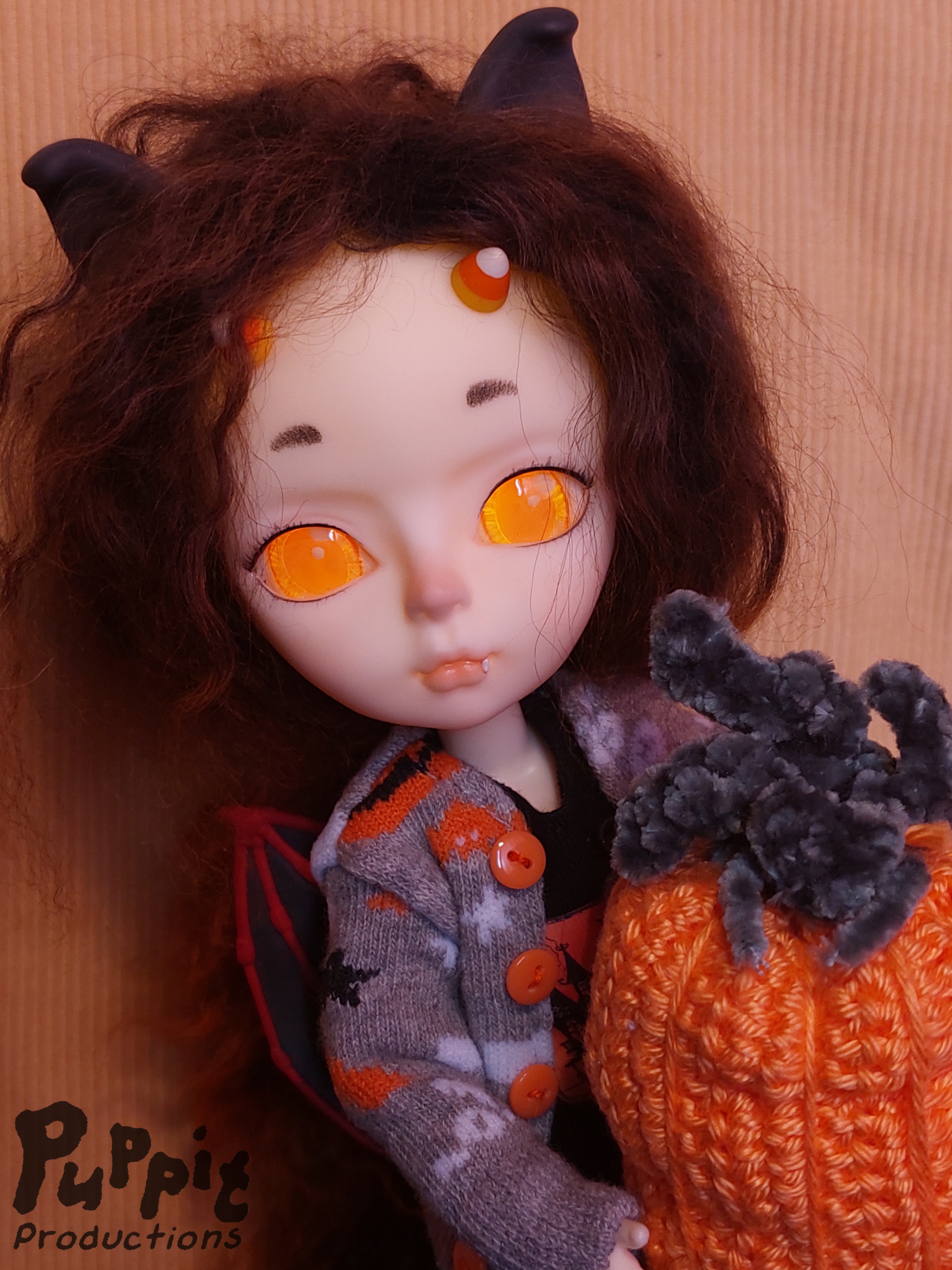 A photograph of a Hujoo Yomi doll with bat ears, candy corn horns, wild wavy black & red hair and glowing orange eyes, wearing a grey sweater with black and orange bats, holding a crocheted pumpkin.