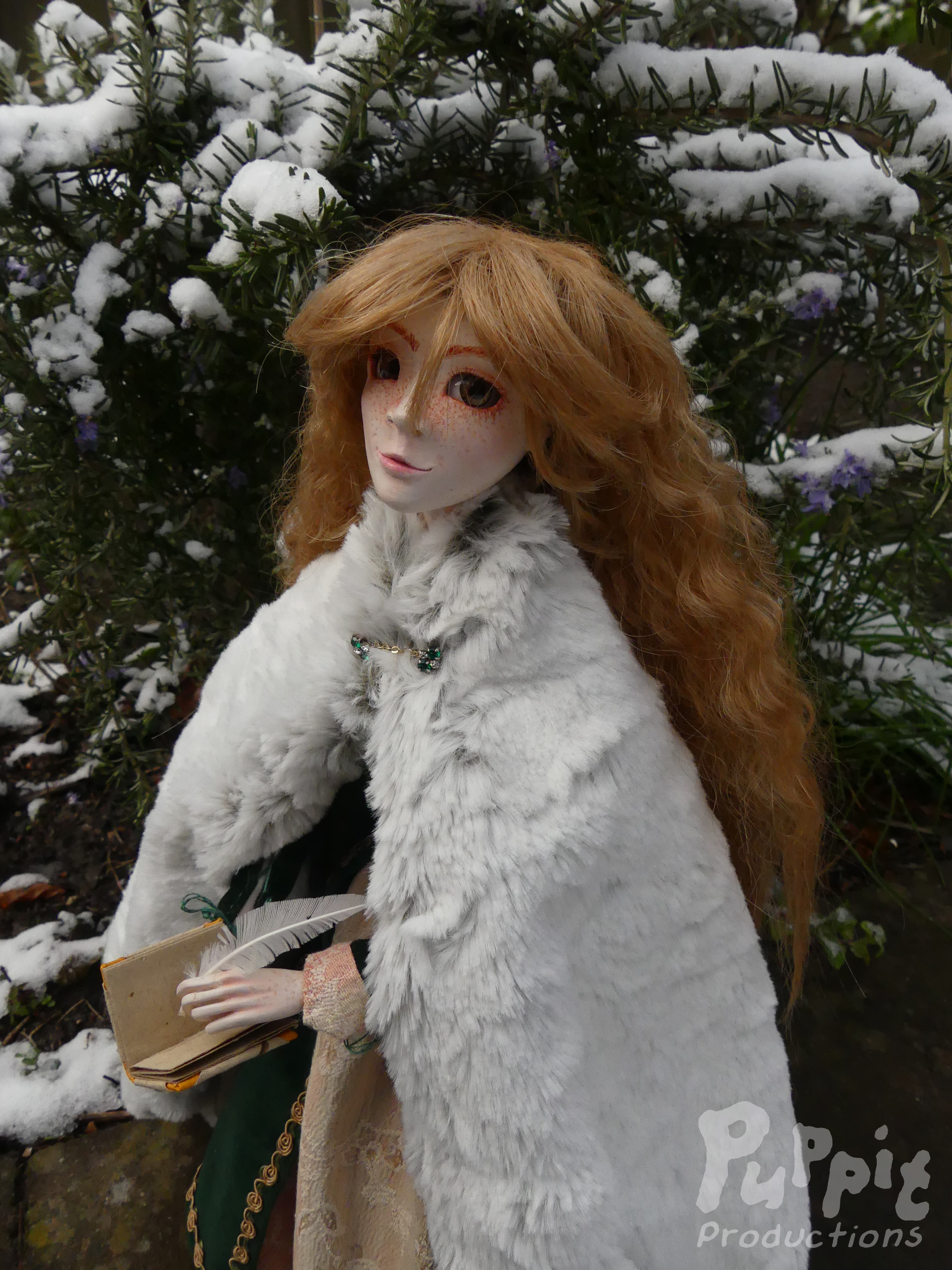  A picture of Ala, a skinny, freckled ball jointed doll with green eyes and long wavy light brown hair, standing in front of a snowy bush, wearing a green Renaissance dress with a white fur cloak. She is holding a notebook and quill and looking at the viewer.